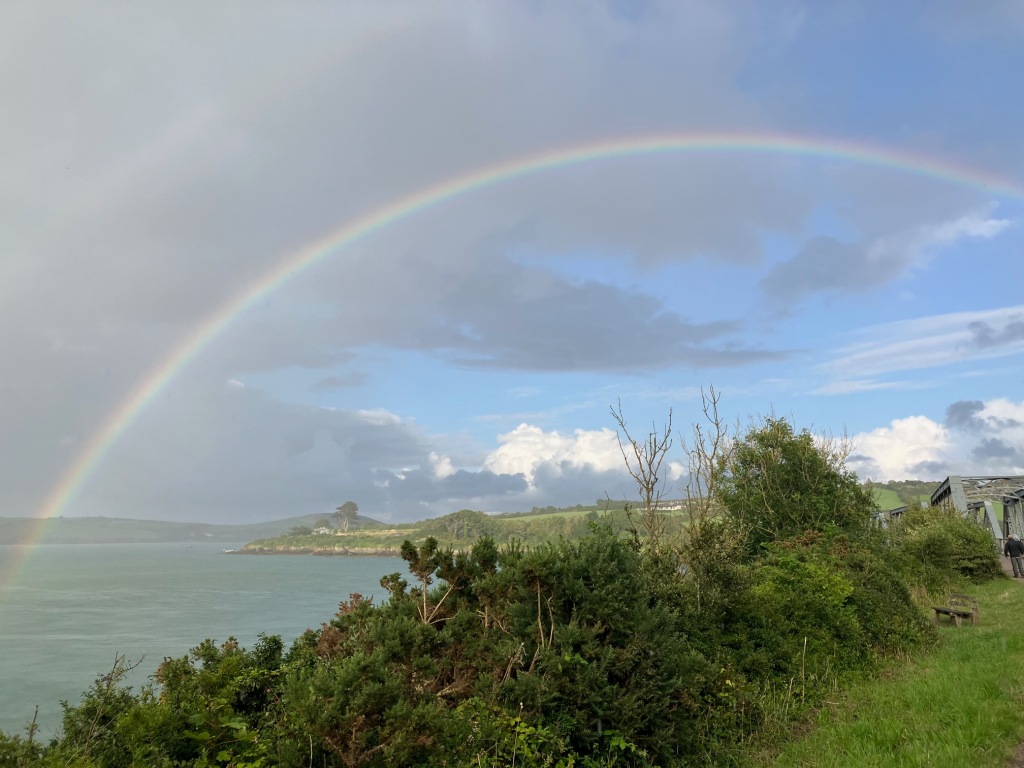 Image of a rainbow extending from the water in the estuary over a metal railway bridge to the right of the photograph. In the foreground underneath the rainbow are some shrubs, and behind it is the coastal landscape. The sky above is blue with large areas of grey cloud.